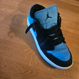 Nike Air Jordan 1 Low blå/svart - Snygga Nike Air Jordan 1 Low i blått och svart med vit sula. Skorna har klassisk snörning, rund tå och ikoniska Jumpman-loggan på plösen. Perfekta för dig som vill sticka ut med stilrena sneakers i skinn och syntet.