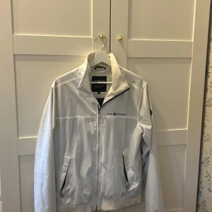 Vit vindjacka från Sail Racing - Snygg vit vindjacka från Sail Racing med dragkedja framtill och två fickor med svarta detaljer. Jackan har hög krage och logga på bröstet, perfekt för dig som gillar sportig och clean stil.