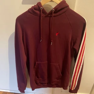 Vinröd hoodie med ränder på ärmen - Vinröd hoodie med huva och snörning, samt en ficka framtill. Ena ärmen har vita, röda och gula ränder längs hela armen. Liten röd broderad logga på bröstet. Perfekt för en chill och sportig look.