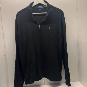 Svart half zip tröja Polo Ralph Lauren - Svart långärmad tröja från Polo Ralph Lauren med half zip och klassisk broderad logga på bröstet. Tröjan har en ribbad struktur och hög krage, perfekt för en clean och stilren look. Passar dig som gillar tidlös design och premiumkänsla.