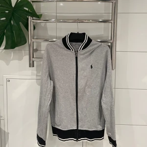 Grå cardigan från Polo - Snygg grå cardigan från Polo med dragkedja och svartvit randig krage. Den har långa ärmar och ribbade muddar i svart och vitt. Perfekt för en avslappnad stil.