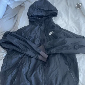 Svart Nike vindjacka med huva - Svart vindjacka från Nike med huva och dragkedja framtill. Jackan har två fickor med dragkedja och ribbade muddar vid ärmsluten. Perfekt för blåsiga dagar och enkel att slänga på sig över en hoodie. Storlek S, ny kostar 1000kr