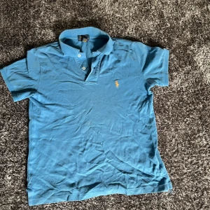 Blå Polo Ralph Lauren t-shirt S - Snygg blå t-shirt från Polo Ralph Lauren i klassisk pikéstil med krage och två knappar framtill. Ikonisk broderad logga i orange på bröstet. Tillverkad i 100% bomull, perfekt för dig som gillar stilren och sportig vibe.