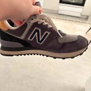 Grå New Balance 574 sneakers - Snygga New Balance 574 sneakers i grått och lila med mesh och mockadetaljer. Klassisk N-logga på sidan, vita snören och bekväm platt sula. Perfekta för en sportig och avslappnad stil. Storlek 37,5