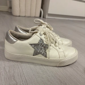 Vita sneakers med glittrig stjärna - Snygga vita sneakers med en glittrig silverstjärna på sidan och silvriga detaljer vid hälen. Skorna har snörning och en platt sula, perfekta för att ge din outfit lite extra bling. 
