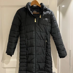 Svart lång ridjacka  - Lång svart pufferjacka från Active RidingWear med huva, dragkedja och snygga guldfärgade detaljer. Jackan har praktiska fickor och tryckknappar nedtill, så den går utmärkt att rida i. Perfekt för kalla dagar i stallet! Köparen står får frakt🤍 
