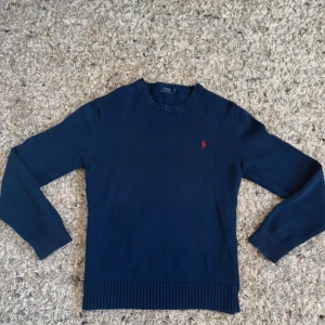 Marinblå stickad tröja Polo Ralph Lauren - Säljer en marinblå stickad tröja från Polo Ralph Lauren i bomull. Klassisk rund halsringning, ribbade muddar och nederkant samt den ikoniska röda loggan broderad på bröstet. Perfekt för dig som gillar stilren och tidlös design.