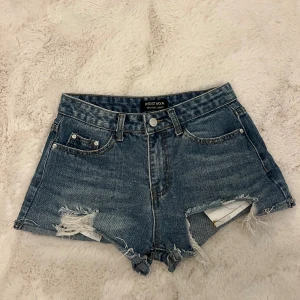 Blå jeansshorts med slitningar Kristhya - Snygga blå jeansshorts från Kristhya med råa kanter och slitna detaljer framtill och på ena bakfickan. Klassisk femficksmodell med bälteshällor och knappstängning. Perfekta för en avslappnad och trendig look.