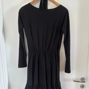 Svart långärmad klänning Cinamoon XS/S - Svart långärmad klänning från Cinamoon i storlek XS/S. Klänningen har markerad midja med resår, volangdetalj nertill och en enkel, stilren design. Tillverkad i mjukt material som ger en skön passform. Perfekt för dig som gillar minimalistisk stil.