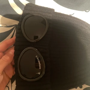 Svart C.P. Company goggle mössa - Svart ribbstickad mössa från C.P. Company med ikoniska goggle-detaljer framtill. Snygg och unik design som sticker ut, perfekt för dig som gillar streetwear. Materialet är mjukt och värmande, och loggan syns tydligt på ena glaset.