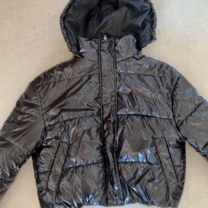 Svart pufferjacka  -  svart, glansig pufferjacka från Boohoo med huva och dragkedja framtill. Jackan har quiltad design, långa ärmar och två fickor på sidorna. Perfekt för dig som vill ha en snygg och varm look under höst och vinter.