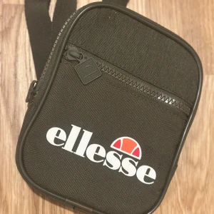 Svart axelväska från Ellesse - Snygg svart axelväska från Ellesse med justerbar rem och stort vitt logotryck framtill. Väskan har dragkedja upptill, ett ytterfack med dragkedja och är tillverkad i slitstark polyester. Perfekt storlek för mobil, plånbok och andra småprylar.