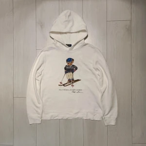 Polo Bear by Ralph Lauren hoodie - Säljer en vit hoodie från Ralph Lauren med det ikoniska Polo Bear-motivet på framsidan. Tröjan har en mysig huva och långa ärmar, perfekt för kyliga dagar. Den är både stilren och lekfull med en skön passform.