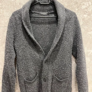 Grå stickad kofta från Dressmann - Mysig grå stickad kofta från Dressmann med sjalkrage och två framfickor. Koftan har knäppning framtill med stora knappar och är långärmad. Perfekt för lager-på-lager och chill höst- och vinterstil.