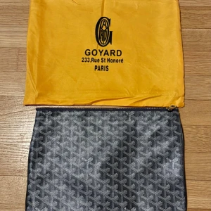 Grå Goy4rd handväska - Säljer en svart kuvertväska från Goy4rd med deras ikoniska grafiska mönster. Väskan har dragkedja upptill och är tillverkad i skinn med detaljer i textil. Perfekt för att bära det viktigaste och ger en lyxig känsla till din outfit.