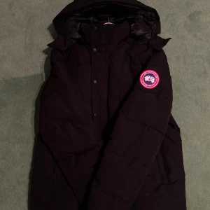 Svart dunjacka från Canada Goose - Säljer en svart dunjacka från Canada Goose med huva och klassisk logga på ärmen. Jackan har quiltad design, tryckknappar framtill och är fodrad med glansigt material. Perfekt för kalla dagar och har en riktigt clean look. Storlek M Finns i Stockholm. Först till kvarn 