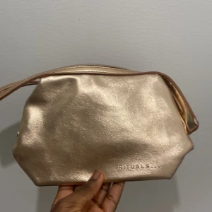 Guldig handväska från Rituals - Snygg guldig från Rituals med stilren design och metallisk finish. Väskan har en praktisk dragkedja upptill och är tillverkad i syntetmaterial med en mjuk, glansig yta. Perfekt storlek för att bära med sig det viktigaste och har ett diskret Rituals-tryck på framsidan. Och kom ihåg att det är för att lägga smink där i massa annat 