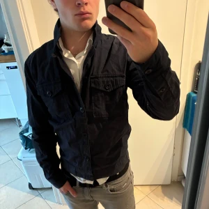 Mörkblå overshirt från Albam - Säljer en mörkblå overshirt från Albam med klassisk krage och två bröstfickor med knappar. Skjortan är i bomull och har en clean, stilren look med lång ärm och knappstängning framtill. Perfekt att slänga över en t-shirt eller skjorta för en snygg layering. Bra skick