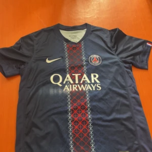 PSG matchtröja Nike blå röd - Paris Saint-Germain fotbollströja från Nike i marinblått med röd och vit grafisk rand längs mitten. PSG-logga på bröstet och Qatar Airways-tryck. Kortärmad, rund hals och tillverkad i lätt polyester. Perfekt för dig som vill representera PSG på träning eller match.