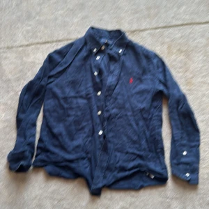 Blå linneskjorta från Ralph Lauren M - Snygg mörkblå linneskjorta från Ralph Lauren i storlek 10-12 år. Klassisk design med knappar framtill och lång ärm. Skjortan har den ikoniska röda Polo-loggan broderad på bröstet och är tillverkad i linne..