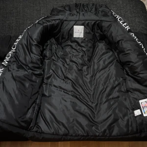 Svart Moncler pufferjacka med logga  - Svart pufferjacka från Moncler med stor logotyp på kragen och klassisk Moncler-patch på ärmen. Jackan har dragkedja framtill, två sidofickor med dragkedja och hög krage. Materialet är glansigt och känns riktigt lyxigt.Stlrk L men passar M bättre