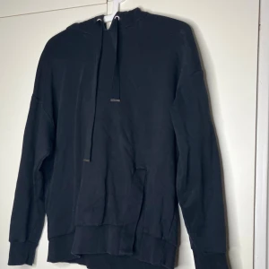 Svart hoodie med huva och snörning - En svart hoodie med huva och långa ärmar. Tröjan har snörning i halsen och ribbade muddar vid ärmslut och nederkant. Perfekt för en chill och avslappnad stil. Gjord i mjuk bomull för extra komfort.