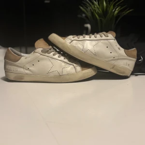 Vita Golden Goose sneakers med beige detaljer - Snygga vita golden goose sneakers med beige mockadetaljer på häl och tå. Skorna har perforerat stjärnmönster på sidorna och klassisk snörning. Tillverkade i skinn med platt sula och rund tå. Perfekta för en avslappnad och trendig look. Grisch, Laidback! Priset är ej hugget i sten!