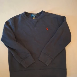 Marinblå sweatshirt från Polo Ralph Lauren - Snygg marinblå sweatshirt från Polo Ralph Lauren med klassisk röd broderad logga på bröstet. Tröjan har rund hals, ribbade muddar och är långärmad. Perfekt för dig som gillar stilrena och tidlösa plagg.