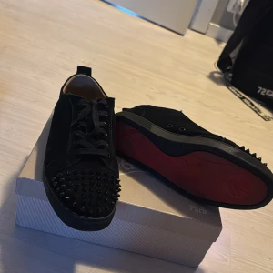 Christian Louboutin  - STORLEK 45.. Säljer ett par svarta sneakers från Christian Louboutin med ikonisk röd sula och svarta nitar på tån. Skorna har snörning och är tillverkade i mocka med rund tå. Kommer med originalkartong och dustbag. Perfekt för dig som vill sticka ut med lyxig streetstyle. A Xopia*