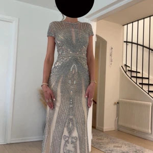 Glittrig långklänning med pärlor - En superfin långklänning i champagnefärgad ton med glittriga silverdetaljer och pärlor. Klänningen har korta ärmar, rund halsringning och ett unikt mönster framtill. Perfekt för bal eller nyår, med en elegant och lyxig känsla.