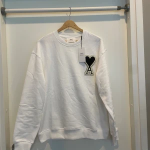 Vit sweatshirt från Ami paris med hjärtdetalj - Vit sweatshirt från Ami Alexandre Mattiussi med stor svart hjärtdetalj och bokstaven A broderad på bröstet. Tröjan har rund hals, ribbade muddar och är tillverkad i mjuk bomull. Perfekt för dig som gillar stilrena och coola detaljer.