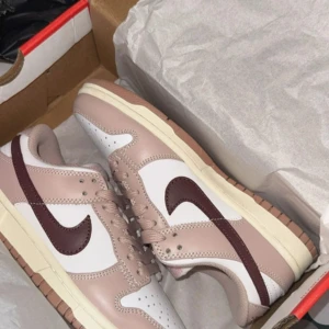 Nike Dunk Low beige brun - Nike Dunk Low sneakers i beige och vitt med brun swoosh. Låg modell med platt sula och rund tå. Ovandel i skinn med perforerade detaljer på tån och klassisk snörning. Snygg färgkombo som lyfter din outfit och passar perfekt till streetwear.