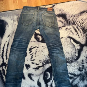 Blå Lee jeans med slim passform - Säljer ett par blå jeans från Lee med snyggt slitna detaljer och klassisk femficksdesign. Jeansen har låg midja och smal passform som sitter tajt längs benen. Materialet är jeans i bomull och färgen är en tvättad blå nyans med ljusare partier. Storlek 31/32