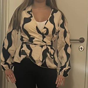 Beige omlottblus med svart mönster - Säljer en beige omlottblus med svarta vågiga mönster. Blusen har långa ärmar, v-ringning och knyt i midjan för en snygg siluett. Perfekt för dig som gillar unika prints och vill sticka ut lite extra.