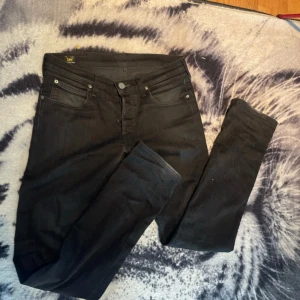 Svarta jeans från Lee - Säljer ett par svarta jeans från Lee med klassisk femficksdesign och smal passform. Jeansen har normal midja och raka ben, tillverkade i mjukt bomullsmaterial med lite stretch för extra komfort. Perfekta för dig som gillar stilrena och enkla jeans. Storlek 31/32