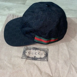 Svart Gucci keps med GG-mönster - Svart keps från Gucci med diskret GG-monogram över hela ytan och klassiska gröna och röda ränder på sidan. Justerbar rem bak i svart skinn. Kepsen är tillverkad i bomull och har märkets etikett invändigt. Perfekt för dig som vill ha en lyxig streetstil.