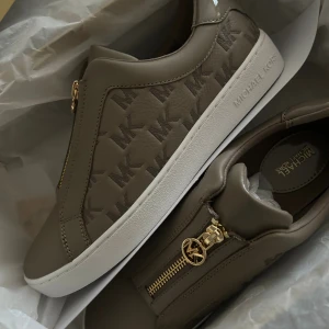 Michael Kors sneakers med gulddragkedja - Snygga sneakers från Michael Kors i beige med MK-logga över hela skon. Skorna har guldfärgade dragkedjor med MK-detalj och vit platt sula. Tillverkade i skinn och syntet, dem är helt oanvända, har ej tagit på mig dem, boxen finns med och kvittot ☺️ det är ett helt nytt skick inga tecken på användning och varan är i orginalförpacking. 