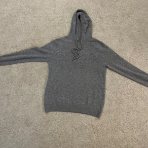 Merino Hoodie - Mysig grå hoodie i stickat material med huva och dragsnören. Tröjan har långärmade ärmar och ribbade muddar vid ärmslut och nederkant. Perfekt för kyliga dagar när du vill vara både bekväm och stilren.