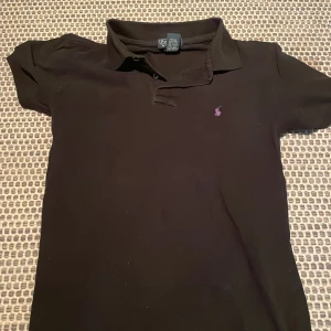 Svart pikétröja från Ralph Lauren - Klassisk svart pikétröja från Ralph Lauren med lila broderad logga på bröstet. Tröjan har korta ärmar, krage och knäppning framtill. Tillverkad i 100% bomull för en skön och luftig känsla. Perfekt för en stilren och avslappnad look.