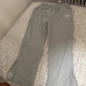 Flared Grå mjukisbyxor - Grå mjukisbyxor med elastisk midja och vita loggor på höften och vid benslutet. Byxorna har raka ben och är tillverkade i mjukt material, perfekta för en avslappnad stil.