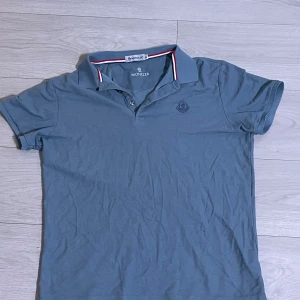 Blå pikétröja från Moncler XL - Snygg blå pikétröja från Moncler i klassisk modell med krage och knappar. Tröjan har broderad logga på bröstet och rödvit detalj på insidan av kragen. Perfekt för dig som gillar stilrena och exklusiva plagg.