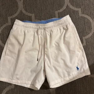Badshorts Polo Ralph Lauren S - Snygga vita badshorts från Polo Ralph Lauren med blå logga broderad på benet. Shortsen har elastisk midja med snörning, innerbyxa i mesh och en bakficka med knapp. Perfekta för stranden eller poolen, klassisk och clean look. Storlek S och använda max 5 gånger.