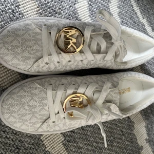 Vita sneakers från Michael Kors - Snygga vita sneakers från Michael Kors med diskret MK-mönster över hela skon. Skorna har rund tå, platt sula och vita snören. På ovansidan sitter en stor guldfärgad MK-logga och baktill en liten guldfärgad detalj. Materialet är syntet med textil känsla.