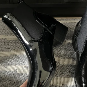Svarta lackade chelsea boots tål regn  - Säljer ett par svarta chelsea boots i glansigt lackmaterial med elastiska paneler på sidorna och spetsig tå. Skorna har en låg klack och är perfekta för dig som vill ha en stilren och edgy look. Passar till både jeans och kjol.