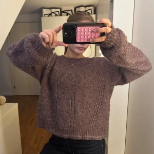 Lila stickad tröja - Mysig lila stickad tröja med snyggt hålmönster framtill. Tröjan har en loose passform, rund halsringning och långa ärmar med breda muddar. Perfekt skick!