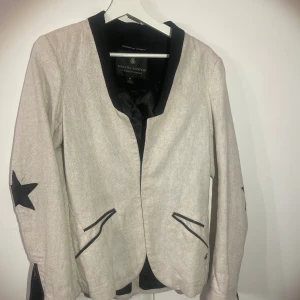 Beige kavaj Maison Scotch med stjärndetaljer - Snygg beige kavaj från Maison Scotch med svarta detaljer, stjärnapplikationer på ärmarna och svarta dragkedjor vid ärmsluten. Jackan har svarta kantband vid fickor och krage, och är tillverkad i ett grovt vävt material som ger en cool och unik look.