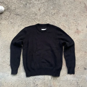 Svart stickad tröja Maison Margiela - Svart stickad tröja från Maison Margiela med klassisk rund halsringning och ribbade muddar. Tröjan har en clean och tidlös look som passar perfekt till jeans eller dressade byxor. Materialet är mjukt och ger en skön känsla hela dagen.