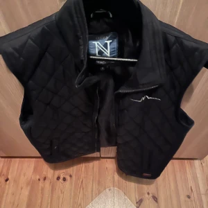 Svart quiltad dunväst från Camden - Quiltad svart dunväst från Camden med hög krage och dragkedja framtill. Västen har ett stilrent rutmönster och en broderad logga på bröstet. Perfekt för lager-på-lager och snygg till både jeans och hoodies.