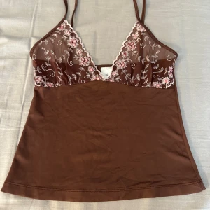 Brun blommig linnetopp med spets - Supersöt brun linnetopp från Tantä med tunna axelband och v-ringad spets upptill. Spetsen är dekorerad med broderade blommor i rosa och vitt, vilket ger en romantisk vibe. Toppen är figurnära och har en mjuk, slät polyesterkänsla. Perfekt att styla med jeans eller kjol.