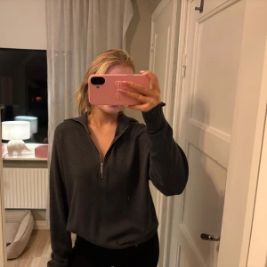 Mörkgrå stickad half zip-tröja - Mysig mörkgrå stickad tröja med half zip-dragkedja och hög krage. Tröjan har långa ärmar med breda muddar och en avslappnad passform. Perfekt för kyliga dagar och enkel att styla till olika outfits.